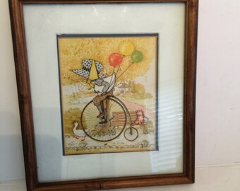 May include: A framed illustration of a child riding a penny-farthing bicycle with balloons attached to the handlebars. The child is wearing a hat and a plaid shirt. A goose and a cat are in the background.