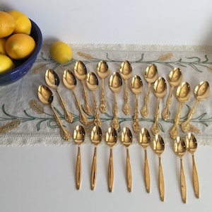 May include: A set of 20 gold-plated teaspoons with ornate designs. The spoons are arranged in two rows on a white tablecloth with a floral pattern.