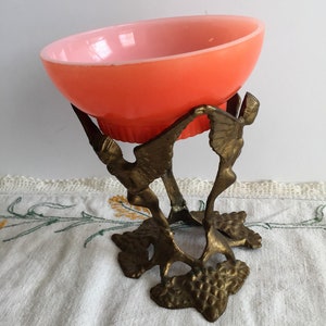 Könnte beinhalten: Eine kleine, rosafarbene Glasschüssel steht auf einem dekorativen, goldenen Metallständer mit Vogelmotiv. Der Ständer hat ein detailliertes, filigranes Design.