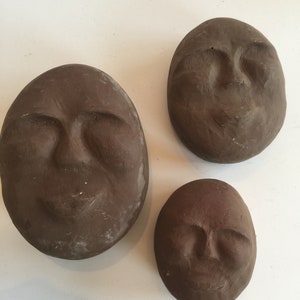 K&ouml;nnte beinhalten: Drei braune Tonskulpturen von Gesichtern mit geschlossenen Augen und einem leichten L&auml;cheln. Die Skulpturen sind oval geformt und haben eine strukturierte Oberfl&auml;che.