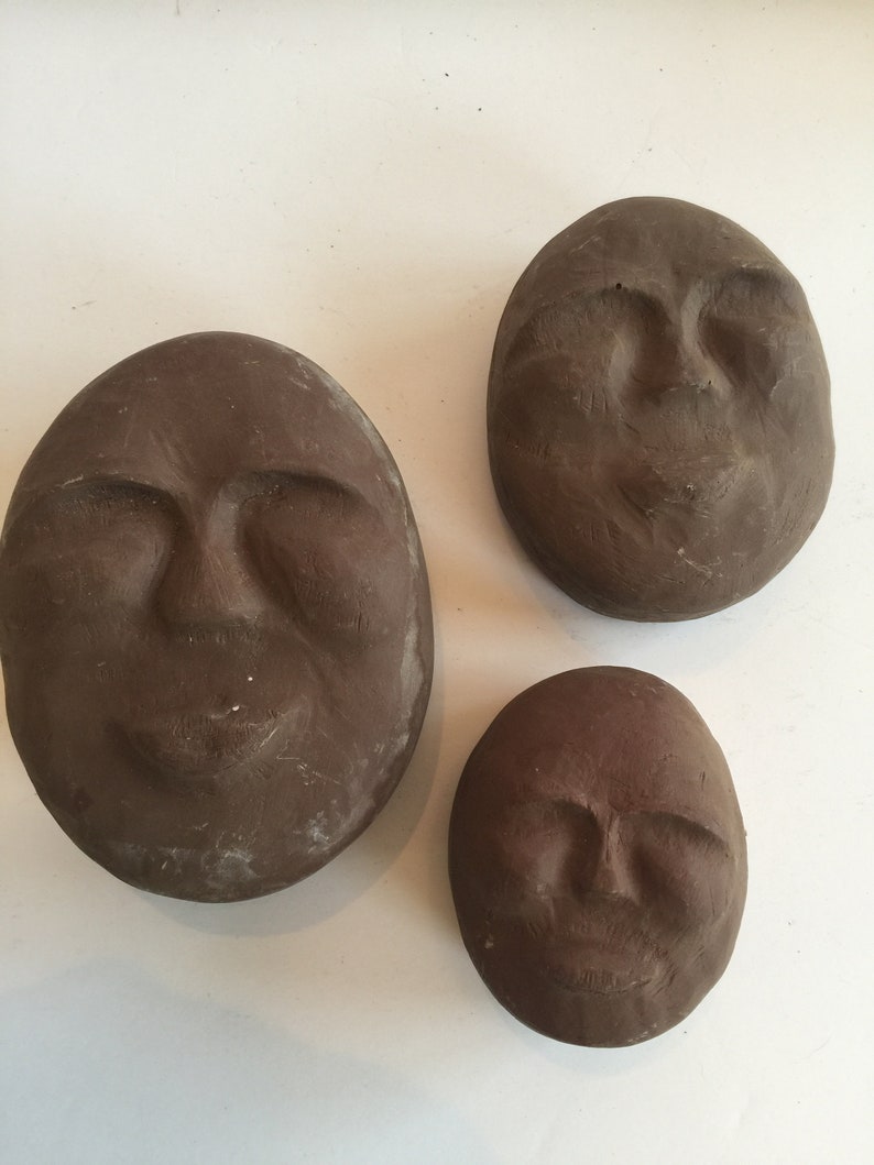K&ouml;nnte beinhalten: Drei braune Tonskulpturen von Gesichtern mit geschlossenen Augen und einem leichten L&auml;cheln. Die Skulpturen sind oval geformt und haben eine strukturierte Oberfl&auml;che.