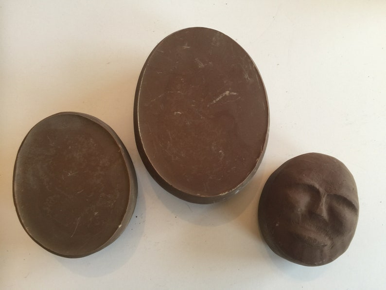 K&ouml;nnte beinhalten: Drei braune Keramik-Wanddekorationen. Zwei sind oval geformt und eine ist ein rundes Gesicht.