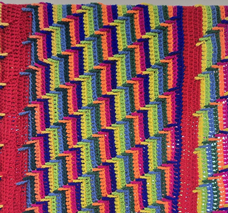 Peut inclure: Une couverture au crochet faite &agrave; la main, aux couleurs vives, avec un motif g&eacute;om&eacute;trique r&eacute;p&eacute;titif. Le design incorpore du rouge, de l'orange, du jaune, du vert, du bleu et du violet, cr&eacute;ant une pi&egrave;ce visuellement frappante et textur&eacute;e. Un &eacute;l&eacute;ment de d&eacute;coration int&eacute;rieure confortable et color&eacute;.