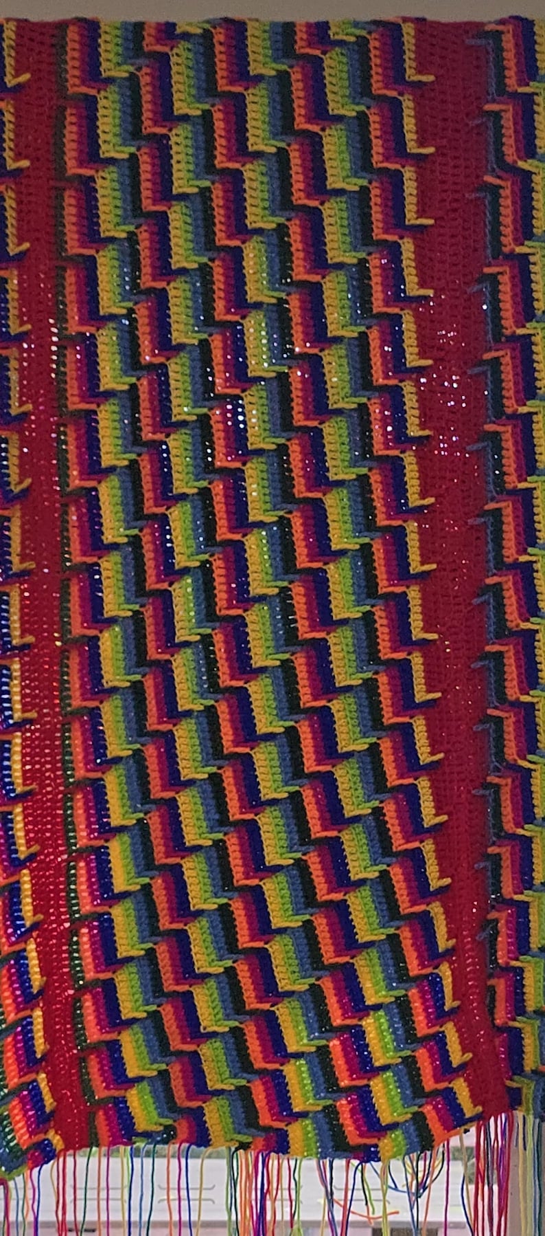 Peut inclure: Une couverture au crochet faite &agrave; la main, aux couleurs vives, avec un motif r&eacute;p&eacute;titif de blocs color&eacute;s arc-en-ciel. La couverture a une bordure et des franges rouges, cr&eacute;ant un textile confortable et accrocheur.