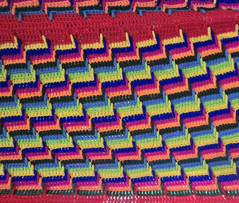 Peut inclure: Une couverture au crochet faite &agrave; la main, aux couleurs vives, avec un motif en chevrons r&eacute;p&eacute;titif. La couverture pr&eacute;sente un spectre de couleurs, notamment le rouge, l'orange, le jaune, le vert, le bleu et le violet, cr&eacute;ant un textile visuellement saisissant et confortable.