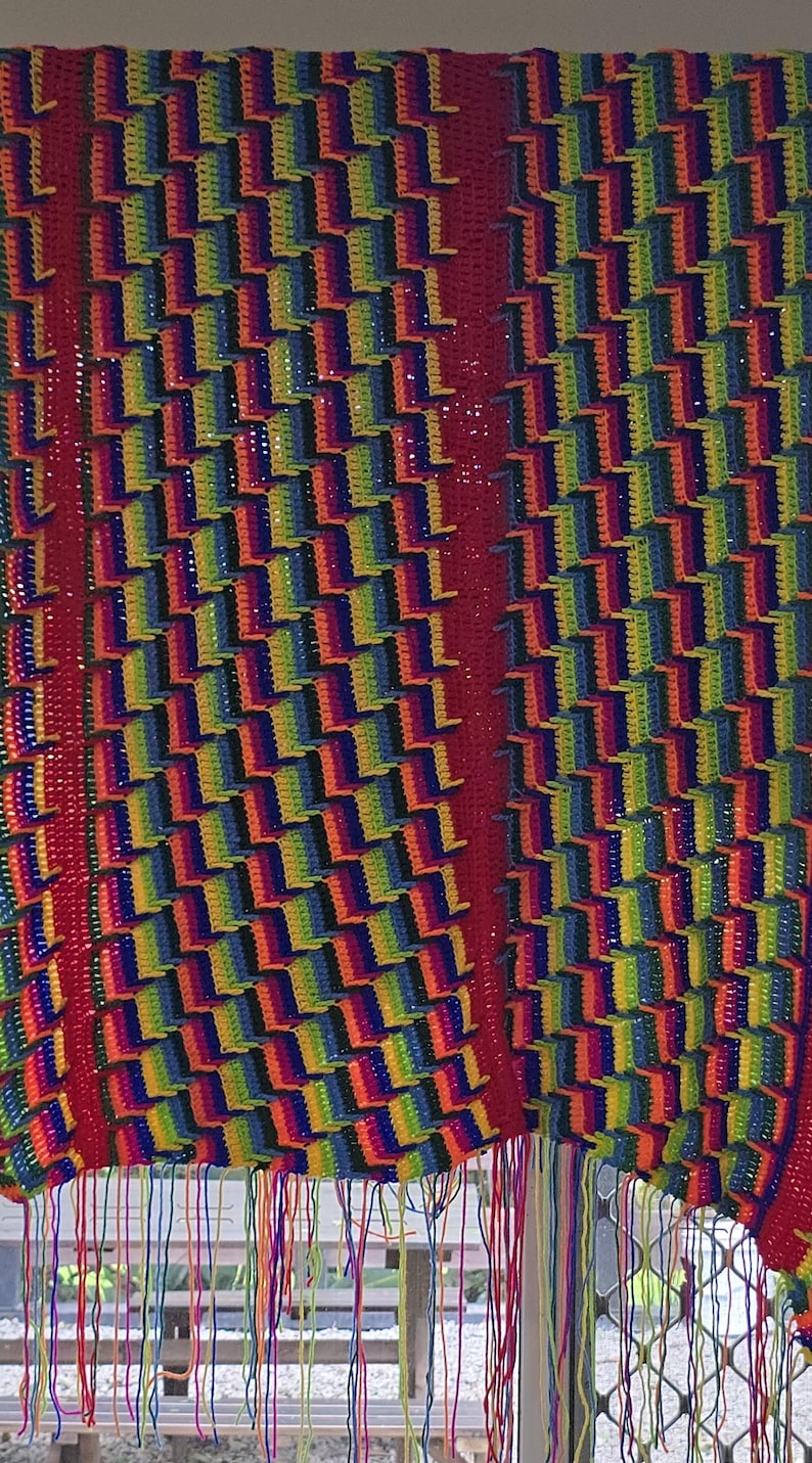 Peut inclure: Un rideau ou tenture murale au crochet fait main et vibrant. Le motif pr&eacute;sente un motif r&eacute;p&eacute;titif de blocs color&eacute;s aux teintes arc-en-ciel, avec une bordure rouge audacieuse et des franges. La pi&egrave;ce est probablement fabriqu&eacute;e &agrave; partir de fil, mettant en valeur un attrait textur&eacute; et artistique.
