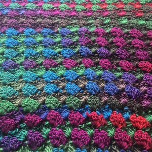 May include: A handmade crochet blanket with a vibrant color palette. The blanket features a repeating pattern of textured stitches in shades of teal, blue, green, purple, and red. The overall design is visually appealing and suitable for home decor.