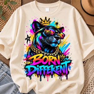 Op de afbeelding: Een crèmekleurig t-shirt met een afbeelding van een zwarte panter die een zonnebril en een gouden ketting draagt, bekroond met een kroon. Het ontwerp bevat levendige verfspatten en de tekst BORN DIFFEKENT in een kleurrijk, druipend lettertype.