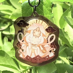 May include: A clear acrylic charm depicting a cartoon character with a halo and flowing robes. The charm has a dark brown border with cloud accents and a black clasp. The background is a vibrant green plant.