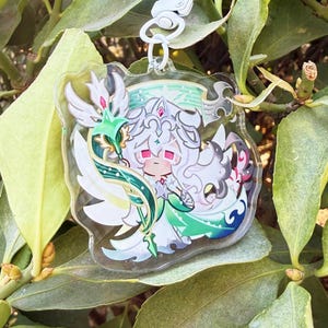 May include: A clear acrylic keychain featuring a cartoon character with white hair, a crown, and pink eyes. The character is surrounded by green and white decorative elements. The keychain has a silver clasp and is set against a backdrop of green leaves.