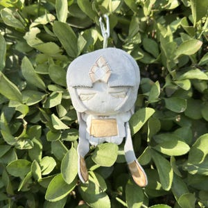 Op de afbeelding: Een lichtgrijze pluche pop met een gestileerd hoofd en lichaam, met een gestileerd gezicht met gesloten ogen en een decoratief hoofddeksel. De pop heeft lange, bungelende aanhangsels en wordt opgehangen aan een wit touwtje. De achtergrond is een dichte opstelling van groene bladeren.