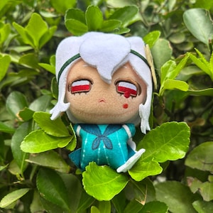 May include: A plush doll with white hair and a teal sailor uniform. The doll has red eyes and a small red mark under one eye. The doll is surrounded by green leaves.
