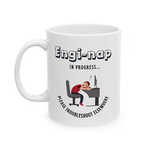 以下が含まれることがあります： 「Engi-nap IN PROGRESS...」の文字と、机で寝ている人の漫画が描かれた白いセラミックマグカップ。「PLEASE TROUBLESHOOT ELSEWHERE」の文字も含まれています。