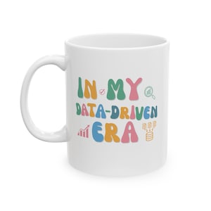 Peut inclure: Mug en céramique blanche avec le texte "IN MY DATA-DRIVEN ERA" en lettres colorées. La tasse présente des icônes liées aux données, notamment une coche, une loupe et un graphique à barres.