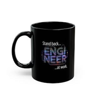 Peut inclure: Mug en céramique noire avec une anse incurvée. Le mug présente le texte "Stand back... ENGINEER ...at work" en blanc et en lettres colorées. Le mug est conçu pour les boissons chaudes.