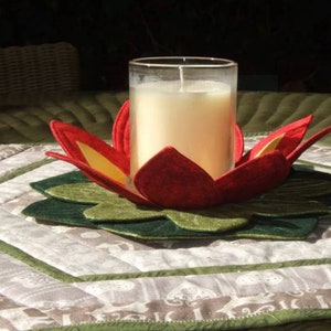 May include: A candle in a clear glass holder sits inside a felt lotus flower. The flower has red petals with yellow accents and green leaves. The candle holder rests on a quilted placemat with a floral pattern.