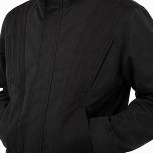 May include: A black jacket with a stand-up collar and vertical stitched details. The jacket has two front pockets and a zipper closure. The sleeves are long and the fabric appears to be a medium weight.