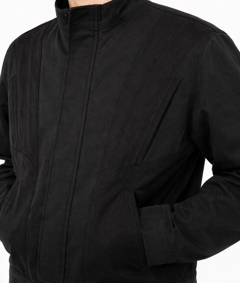 May include: A black jacket with a stand-up collar and vertical stitched details. The jacket has two front pockets and a zipper closure. The sleeves are long and the fabric appears to be a medium weight.