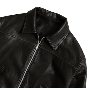 May include: Black leather jacket with a collared neckline and a silver zipper closure. The jacket has a classic design with visible stitching details and a smooth, matte finish. The jacket appears to be new and unworn.