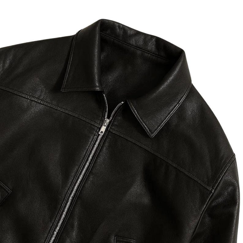 May include: Black leather jacket with a collared neckline and a silver zipper closure. The jacket has a classic design with visible stitching details and a smooth, matte finish. The jacket appears to be new and unworn.