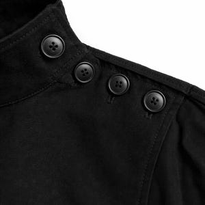 May include: Close-up of a black garment featuring a high collar and three decorative buttons on the shoulder. The buttons are round and dark, contrasting against the fabric. The stitching is visible.