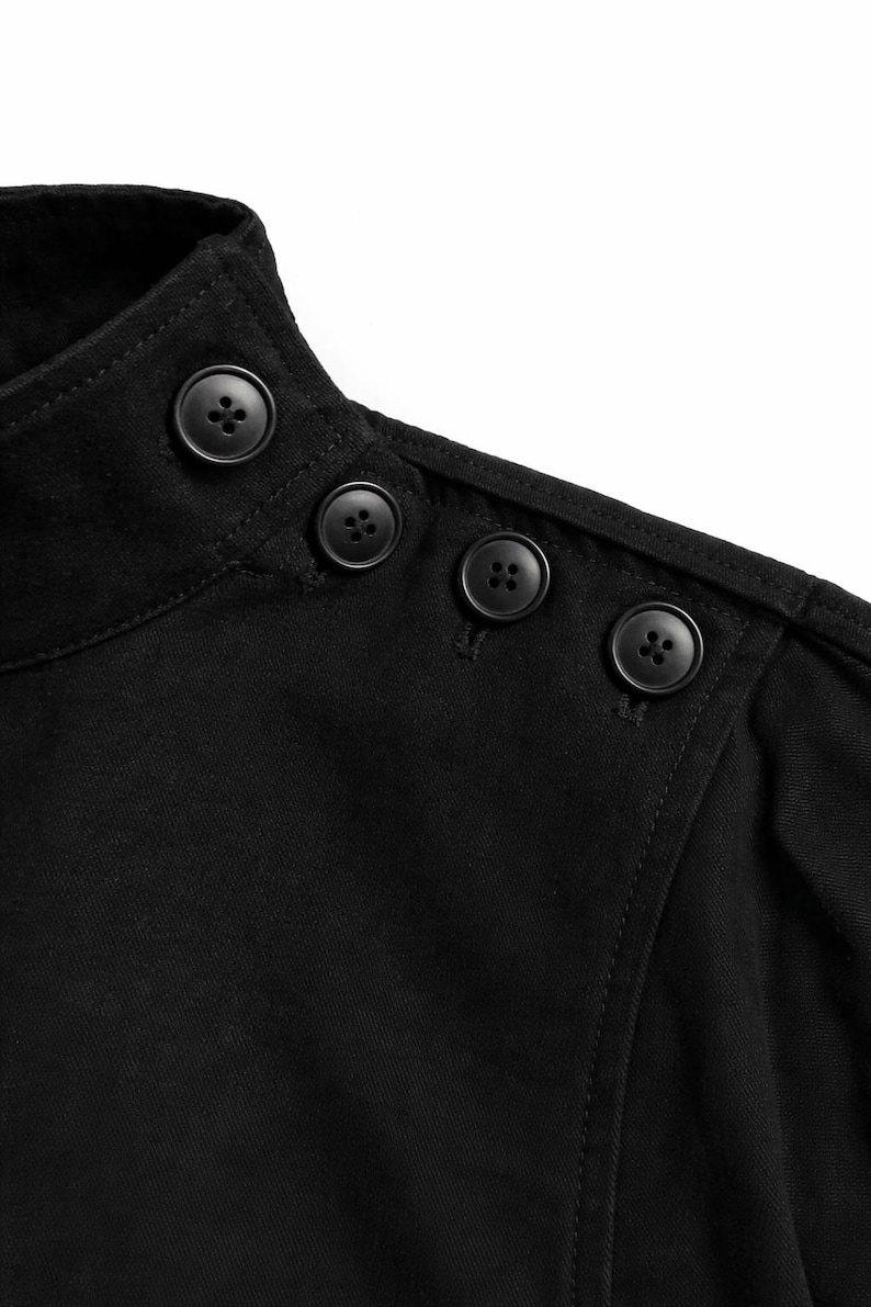 May include: Close-up of a black garment featuring a high collar and three decorative buttons on the shoulder. The buttons are round and dark, contrasting against the fabric. The stitching is visible.