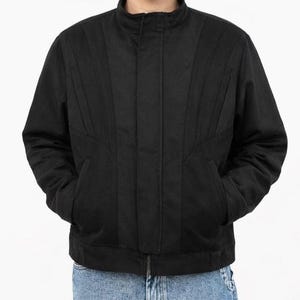 May include: A black jacket with a stand-up collar and vertical stitched details. The jacket has two front pockets and a zipper closure. The model is wearing blue jeans. The jacket is a casual outerwear garment.