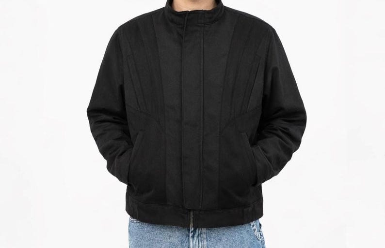 May include: A black jacket with a stand-up collar and vertical stitched details. The jacket has two front pockets and a zipper closure. The model is wearing blue jeans. The jacket is a casual outerwear garment.