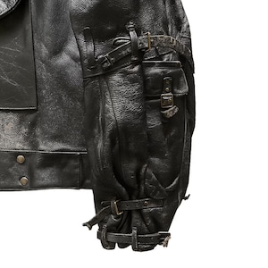 May include: Close-up of a black leather jacket with multiple pockets and straps. The jacket features a textured surface and metal buckles. The sleeve has a gathered design with adjustable straps. The jacket appears to be vintage or distressed.