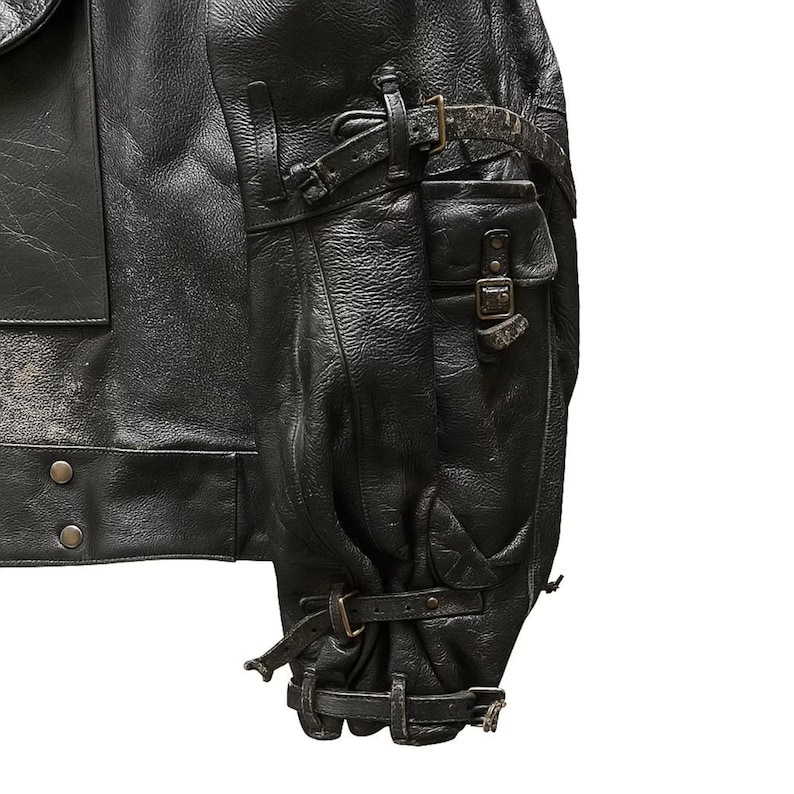 May include: Close-up of a black leather jacket with multiple pockets and straps. The jacket features a textured surface and metal buckles. The sleeve has a gathered design with adjustable straps. The jacket appears to be vintage or distressed.