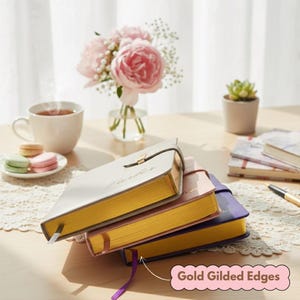 May include: A stack of three journals with gold gilded edges. The journals are in cream, pink, and purple. A gold clasp secures the cream journal. A cup of tea, macarons, and flowers are in the background.