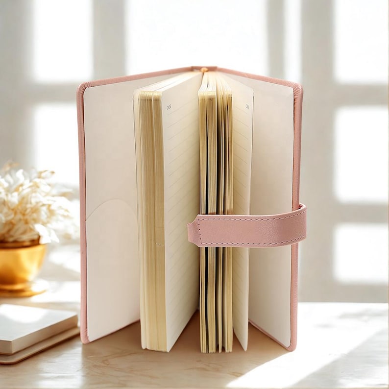 May include: Open pink leather journal with lined pages and a pink strap closure. The journal is open, revealing the cream-colored pages. A small pocket is visible on the inside cover. The journal is on a wooden surface.