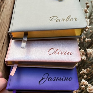 May include: A stack of three personalized notebooks in gray, pink, and purple. Each notebook has a name printed on the cover: Parker, Olivia, and Jasmine. The edges of the notebooks are gold, and each has a ribbon bookmark.