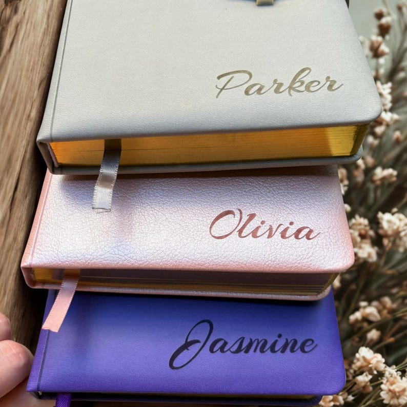 May include: A stack of three personalized notebooks in gray, pink, and purple. Each notebook has a name printed on the cover: Parker, Olivia, and Jasmine. The edges of the notebooks are gold, and each has a ribbon bookmark.