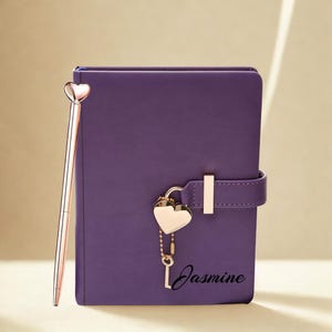 May include: A purple journal with a heart-shaped lock and a matching pen. The journal has a strap closure and the name "Jasmine" printed on the front. The pen and lock are gold-colored.