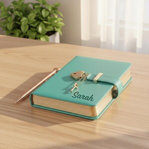 May include: A teal-colored journal with gold-edged pages and a heart-shaped lock. The journal has a gold clasp and a key charm. The name "Sarah" is printed on the cover. A rose gold pen rests beside the journal.