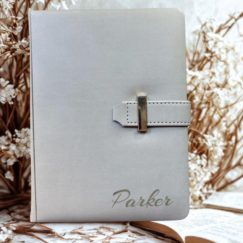 May include: A light gray journal with a silver clasp closure and the name "Parker" engraved on the cover. The journal is rectangular and has a leather-like texture. The background includes dried flowers.
