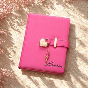 May include: A bright pink journal with a heart-shaped lock and key charm. The journal has a gold-colored clasp and the name "Lauren" printed in cursive on the front. The journal is closed and appears to be made of leather or a similar material.