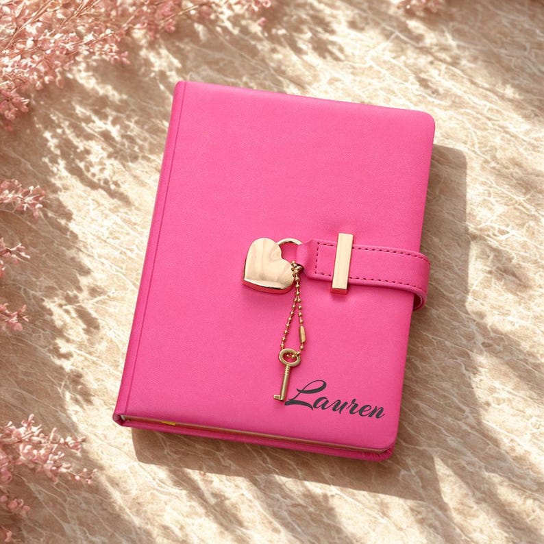 May include: A bright pink journal with a heart-shaped lock and key charm. The journal has a gold-colored clasp and the name "Lauren" printed in cursive on the front. The journal is closed and appears to be made of leather or a similar material.