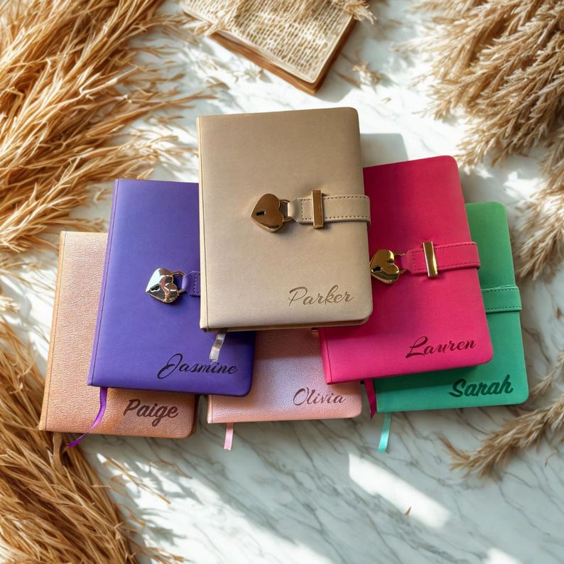 May include: A collection of colorful, personalized journals with heart-shaped lock closures. The journals are in various colors, including tan, purple, pink, and green. Each journal has a name engraved on the front. A small open book is in the background.