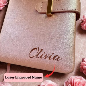 May include: A rose gold leather notebook with the name "Olivia" laser engraved on the cover. The text "Laser Engraved Name" is also visible. The notebook has a gold-colored clasp and a pink ribbon bookmark.