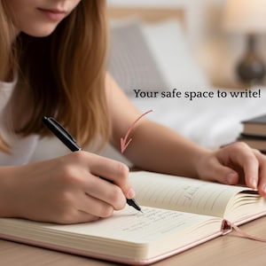 May include: A person writing in a pink journal with a black pen. The journal is open to a blank page with lines. The text "Your safe space to write!" is above the journal. The journal has a pink ribbon bookmark.