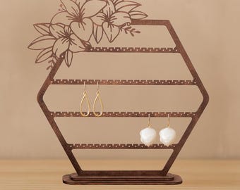 May include: A brown wooden earring holder in a hexagonal shape, adorned with a floral design at the top. The holder features multiple rows for displaying earrings, with two pairs of earrings hanging from it. The earrings are gold and white.