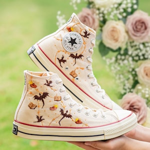 May include: Pair of high-top canvas sneakers in cream with embroidered bats, books, and hats. The sneakers feature a red stripe on the sole and the Converse All Star logo. The shoes are held in front of a blurred floral background.