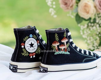 May include: Black high-top sneakers with embroidered mushroom and foliage designs. The sneakers feature white laces, a white rubber sole, and the Converse All Star logo. The design includes red, brown, and green accents.