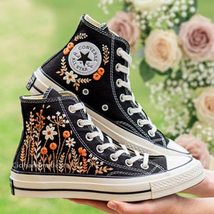 Op de afbeelding: Zwarte hoge sneakers met witte veters en zolen. De sneakers hebben geborduurde bloemmotieven in oranje en wit. Het Converse All Star-logo is zichtbaar. De tekst "Cicihandmade" is zichtbaar op de zijkant van de schoen.