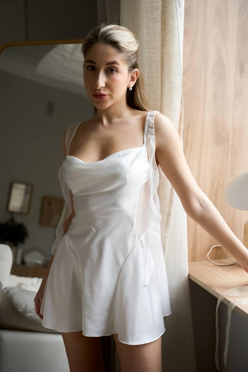 May include: A white satin slip dress with sheer overlay details. The dress features a draped neckline and spaghetti straps. The skirt flares out from the waist. The dress is knee-length and appears to be made of a lightweight fabric.