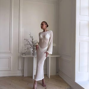 May include: A white, long-sleeved, open-weave maxi dress. The dress has a fitted silhouette and a sheer appearance. The model is wearing a white bikini underneath. The dress has a round neckline and flared sleeves. The model is wearing clear heels.