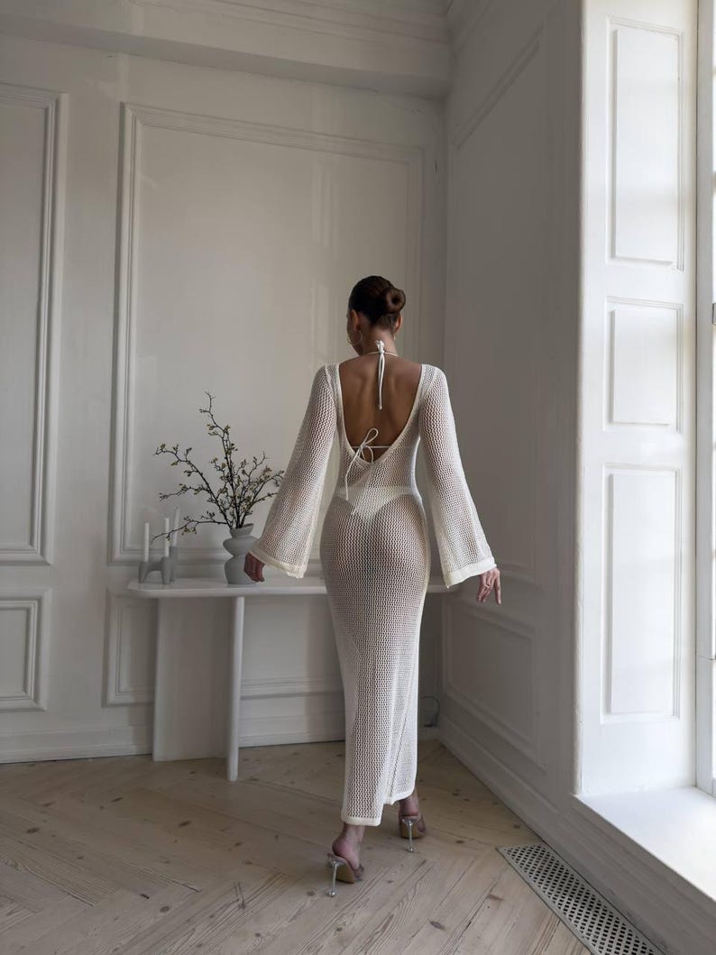 May include: A white, open-weave maxi dress with long, flared sleeves and a low back. The dress is worn with a white swimsuit. The model is wearing clear heels. The dress is a beach cover-up.