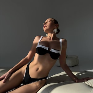 May include: Black and white bikini set with a contrasting white trim. The top features a structured design with white straps, and the bottoms are high-cut. The model is wearing flower-shaped earrings and a gold bracelet.
